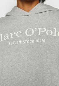 Gros plan d'une personne portant un hoodie gris clair avec un texte blanc indiquant Marc O'Polo Est. à Stockholm sur la poitrine.