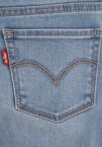 Tasca posteriore dei jeans in denim azzurro chiaro, contornata da cuciture marroni. Presenta un design curvo distintivo e un'etichetta del marchio rossa sul lato.