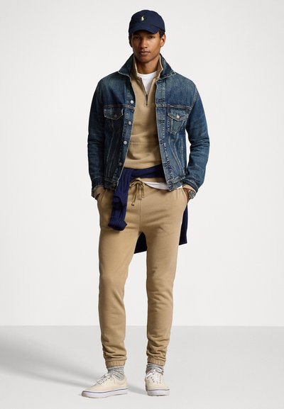 Jeansjacke, beiger Sweatshirt, beiger Jogger, dunkelblauer Pullover um die Taille gewickelt, dunkelblappe Kappe, helle Sneaker, strukturierte Materialien, lässiges Design.