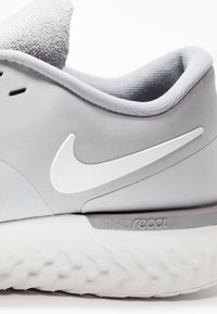 Zapatilla de color gris claro con un material suave, que presenta un logo blanco de Nike y la marca "nike react" en la suela. Detalle texturizado en el cuello.