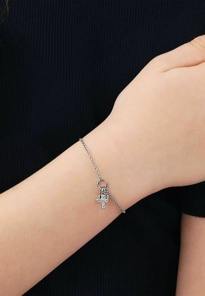 Zarter silberner Gliederarmband am Handgelenk mit kleinen Kreuz- und herzförmigen Anhängern, getragen vor schwarzem Stoffhintergrund.