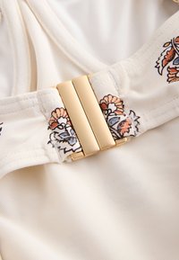 Beiger Stoff mit Blumenstickerei und einem goldfarbenen Metallverschluss. Glatte Textur und ein tailliertes Design für ein strukturiertes Erscheinungsbild.