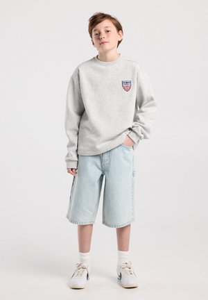 Junge Person steht mit der Hand in der Tasche, trägt einen hellgrauen Pullover mit Aufnäher, hellblaue Jeanskurzshorts bis zum Knie, weiße Socken und weiße Turnschuhe.