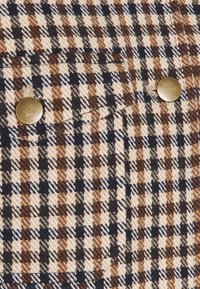 Tela de cuadros en marrón, beige y azul marino con tejido texturizado. Cuenta con un pequeño bolsillo en el pecho y botones de tono dorado.