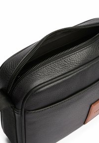 Borsa in pelle nera con superficie testurizzata, chiusura con zip e un'accento in pelle marrone. Presenta un ampio interno e una tracolla piatta.