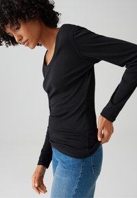 Vrouw met krullend haar die een nauwsluitend zwart shirt met lange mouwen en een blauwe spijkerbroek draagt, terwijl ze de zoom van het shirt tegen een effen achtergrond recht trekt.