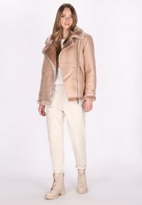 Giacca in pelle beige con colletto di pelliccia, dotata di chiusura con zip e texture morbida. Abbinata a pantaloni di colore chiaro e stivali beige con lacci.