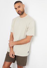 Beige bomulds t-shirt med korte ærmer, rund halsudskæring og lige snit, parret med mørkegrønne shorts. Etiketten er synlig nederst til venstre.