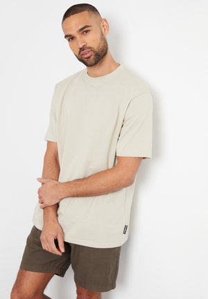 Camiseta de algodón beige con mangas cortas, cuello redondo y corte recto, combinada con pantalones cortos verde oscuro. Etiqueta visible en la parte inferior izquierda.