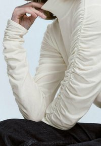 Beige Langarmoberteil mit hohem Kragen und geraffter Textur an den Ärmeln, kombiniert mit dunkler Denim. Nahaufnahme der Handposition.