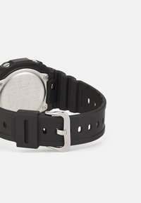 Montre-bracelet en caoutchouc noir avec un bracelet texturé, boucle en métal argenté et un fond en acier inoxydable affichant des détails gravés.