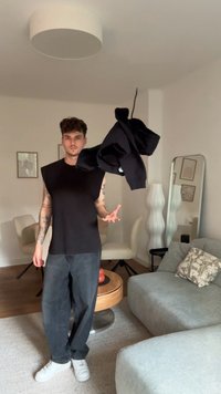 Top preto sem mangas com ombros largos, segurado por uma pessoa. Jeans escuros e ténis brancos. Interior minimalista com mobiliário claro.