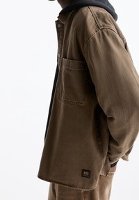 Persoon in een bruine jas over een zwarte hoodie, handen in de zakken, witte achtergrond, gedeeltelijk zijaanzicht.