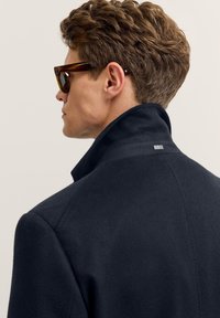 Marineblauer Wollmischungsmantel mit hohem Kragen und kleinem Logo-Detail auf der Rückseite, getragen mit dunklen Sonnenbrillen und zeigt eine glatte Textur.