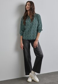 Grön mönstrad blus med lösa ärmar, kombinerad med högmidjade mörkgrå jeans och vita sneakers, står mot en grå vägg.