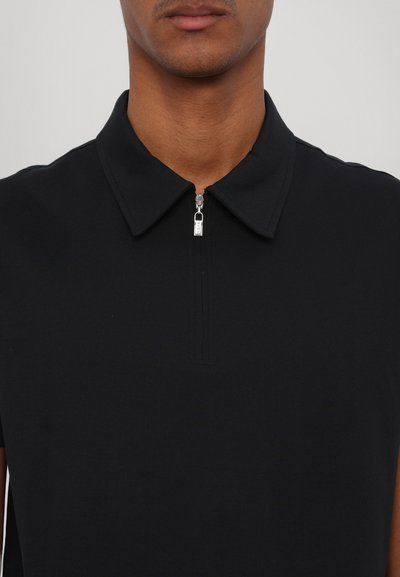 Polo preto com colarinho estruturado e um pequeno fecho de zíper no pescoço, apresentando um tecido texturizado e um design ajustado.