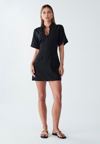 Črna obleka s ovratnikom, kratkimi rokavi in dvema sprednjima žepoma. Gladek material, do kolen, minimalističen dizajn.