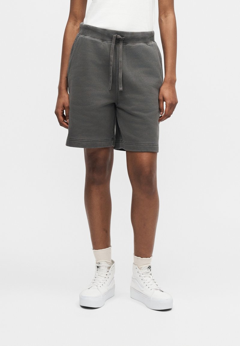 Persona che indossa pantaloncini grigio scuro con coulisse, scarpe da ginnastica alte bianche e calzini chiari su uno sfondo chiaro uniforme.