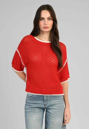 Jeune femme aux longs cheveux foncés portant un pull court rouge en crochet ample par-dessus un jean en denim, debout devant un fond gris uni.