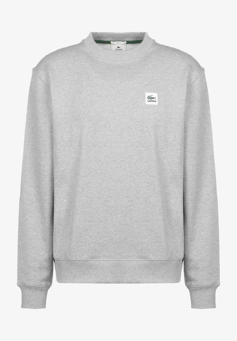 Felpa grigia con maniche lunghe, scollo a girocollo a coste e polsini elastici. Presenta una piccola toppa con il logo Lacoste sul davanti. Texture morbida.