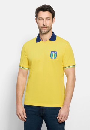 Mann trägt ein gelbes Polohemd mit blauem Kragen und einem Brasilien-Emblem auf der Brust, kombiniert mit dunkelblauen Jeans.