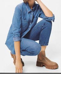 Stivali alla caviglia marroni con suola spessa, realizzati in materiale liscio. Indossati con una camicia di jeans blu e jeans, presentando un look casual a strati.