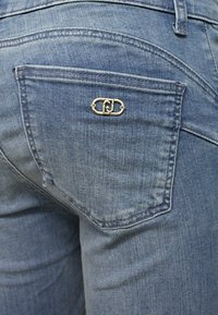 Jeans in denim blu con una tasca posteriore che presenta un dettaglio del logo in tonalità oro. Il materiale presenta una leggera texture con aree sbiadite.