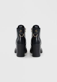 Bottes de cheville en cuir noir avec un haut talon bloc, fermeture éclair et accents en métal doré. Présente un détail de sangle en boucle au niveau du talon.