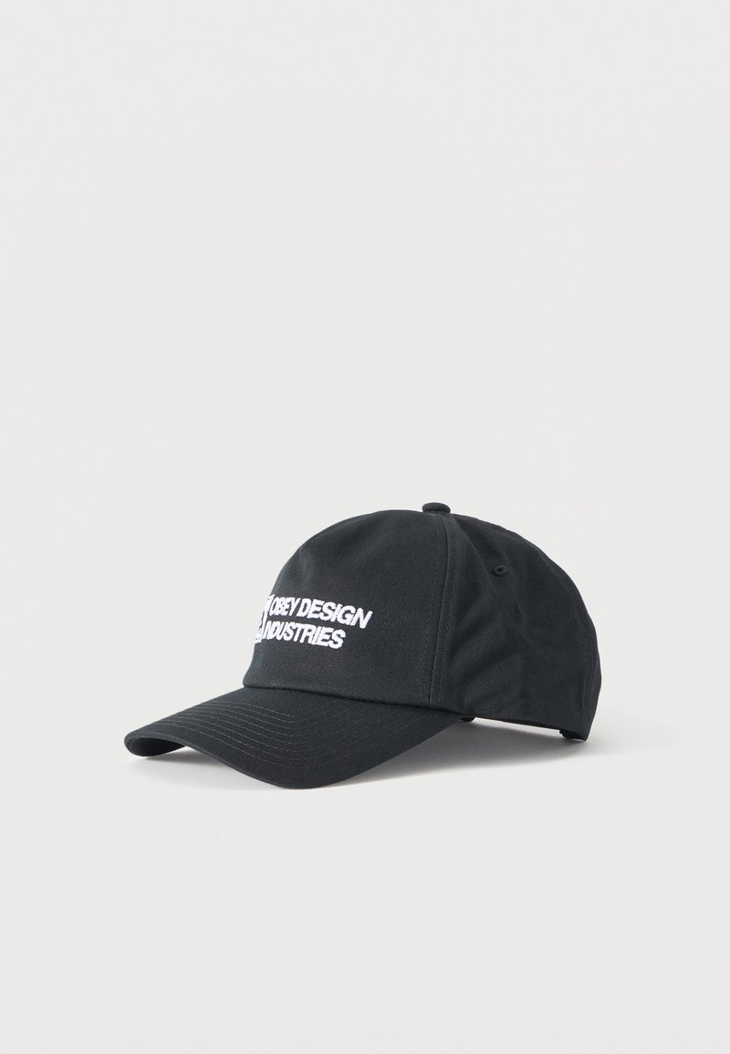 Μαύρο καπέλο μπέιζμπολ με λευκό κεντημένο κείμενο "OBEY DESIGN INDUSTRIES" στο μπροστινό μέρος, εμφανίζεται σε απλό φωτεινό φόντο.