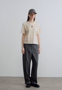 Μπεζ βαμβακερό μπλουζάκι με κοντά μανίκια και μικρό λογότυπο, συνδυασμένο με γκρι παντελόνι με λεπτές ρίγες και γκρι καπέλο, φορεμένο με μαύρα παπούτσια.