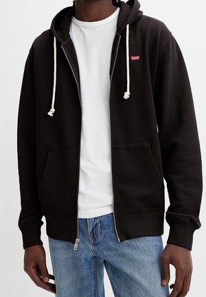 Uomo che indossa una felpa nera con zip, aperta, con cordini bianchi sopra una t-shirt bianca e jeans blu, in piedi contro uno sfondo semplice.