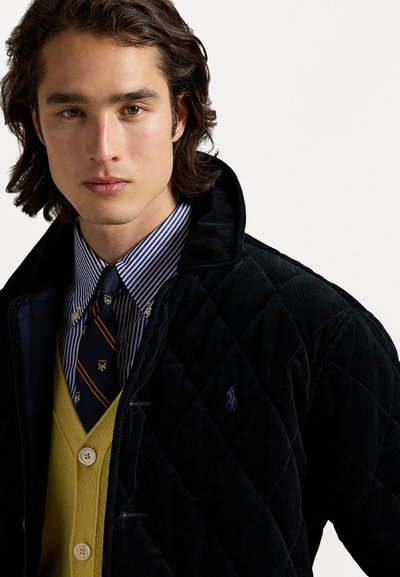 Melna šņorēta kvilts jakas ar augstu apkakli, kas uzvilkta virs dzeltenā kardigāna un zilo un baltu svītraino krekli ar rakstainu kaklasaiti.