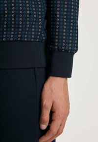 Schwarzer Sweatshirt mit blauen und weißen kreisförmigen Mustern, mit einem gerippten Bund. Eine Hand ruht in der Nähe einer passenden schwarzen Hose.