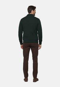 Suéter verde de punto con cuello de canalé, dobladillo ajustado y mangas largas, combinado con pantalones marrones y zapatos marrones. La vista es desde la parte posterior.