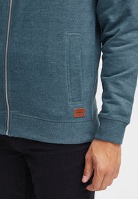 Haut à capuche zippé bleu sarcelle en tissu doux avec un ourlet et des poignets côtelés. Dispose d'une poche latérale et d'un patch en cuir brun à la taille.