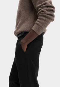 Brun ribbet sweater båret med sorte bukser. Sweateren har en afslappet pasform og opsmøgede ærmer; bukserne har en sidelomme og knapdetaljer.