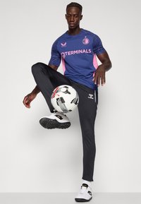 Vêtements de sport pour hommes : maillot marine et rose avec logo, pantalon slim noir, baskets blanches et noires, personne tenant un ballon de foot à motifs.