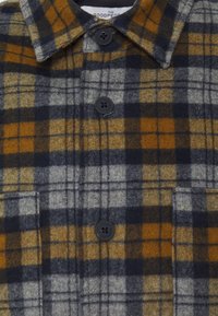 Brown, navy, and gray plaid shirt with a button-down collar, featuring black buttons and two pockets on the front. Made of soft fabric.