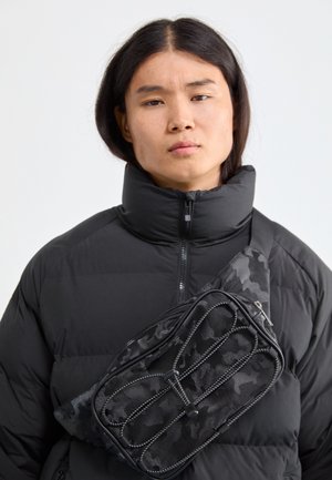 Schwarze Nylon-Bauchtasche mit Camouflage-Muster und Reißverschlusstaschen, diagonal über einer schwarzen Steppjacke mit hohem Kragen getragen.