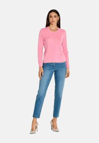Rosa Langarmpullover mit Rundhalsausschnitt, kombiniert mit blauen Jeans. Das Model trägt gemusterte High Heels und präsentiert eine figurbetonte Silhouette.