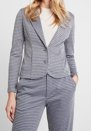 Vrouw draagt een zwart-wit pied-de-poule blazer en bijpassende broek met een lichtgekleurde top, staand met één hand in de zak.