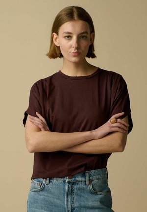 Jeune femme aux cheveux bruns courts, portant un t-shirt marron foncé et un jean bleu, debout les bras croisés sur un fond beige.