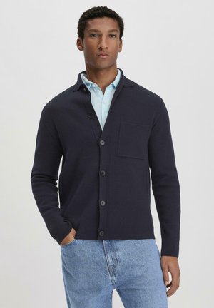 Navy cardigan made of knit fabric with a collar, seven buttons, a chest pocket, and fitted design, worn over a light blue shirt.