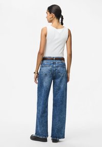 Γυναίκα με πλεγμένα μαλλιά που φοράει λευκό μπλουζάκι χωρίς μανίκια, μπλε παντελόνι φαρδιάς γραμμής με ψηλή μέση, μαύρα παπούτσια και χρυσούς κρίκους σκουλαρίκια, γυρισμένη από την πλάτη.