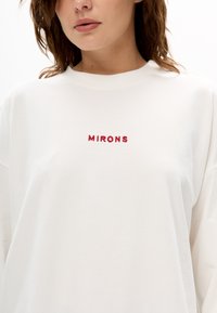 Weißes Langarmhemd aus weichem Stoff mit roter, gestickter Aufschrift "MIRONS" in der Mitte der Brust. Rundhalsausschnitt und lockere Passform.
