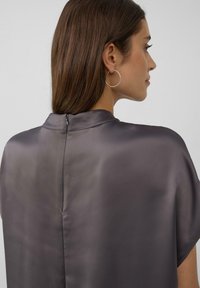 Top en satin gris avec un col haut, des manches courtes et une fermeture éclair discrète à l'arrière. Texture lisse et éclat subtil. Boucles d'oreilles en argent en forme de cercle visibles.