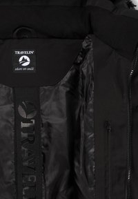 Zwarte jas geopend om de binnenvoering te tonen met het label "TRAVELIN' where we walk" en een verticale gemarkeerde band op het binnenpaneel aan de achterkant.