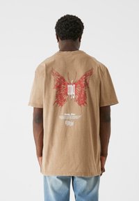 Brown cotton t-shirt featuring a large red butterfly design on the back, with white text underneath. Short sleeves and a relaxed fit.