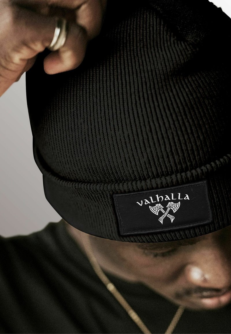 Black knit beanie with a textured pattern, featuring a rectangular black label with the word “valhalla” and axe graphic in white.