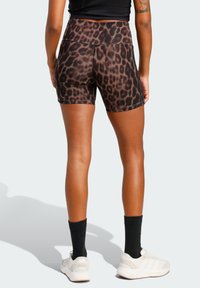 Γυναίκα που φοράει σορτς με υψηλή μέση σε καφέ και μαύρο λεοπάρ μοτίβο, μαύρες κάλτσες, λευκά αθλητικά παπούτσια και μαύρο cropped τοπ, που στέκεται με την πλάτη προς τα εμπρός.